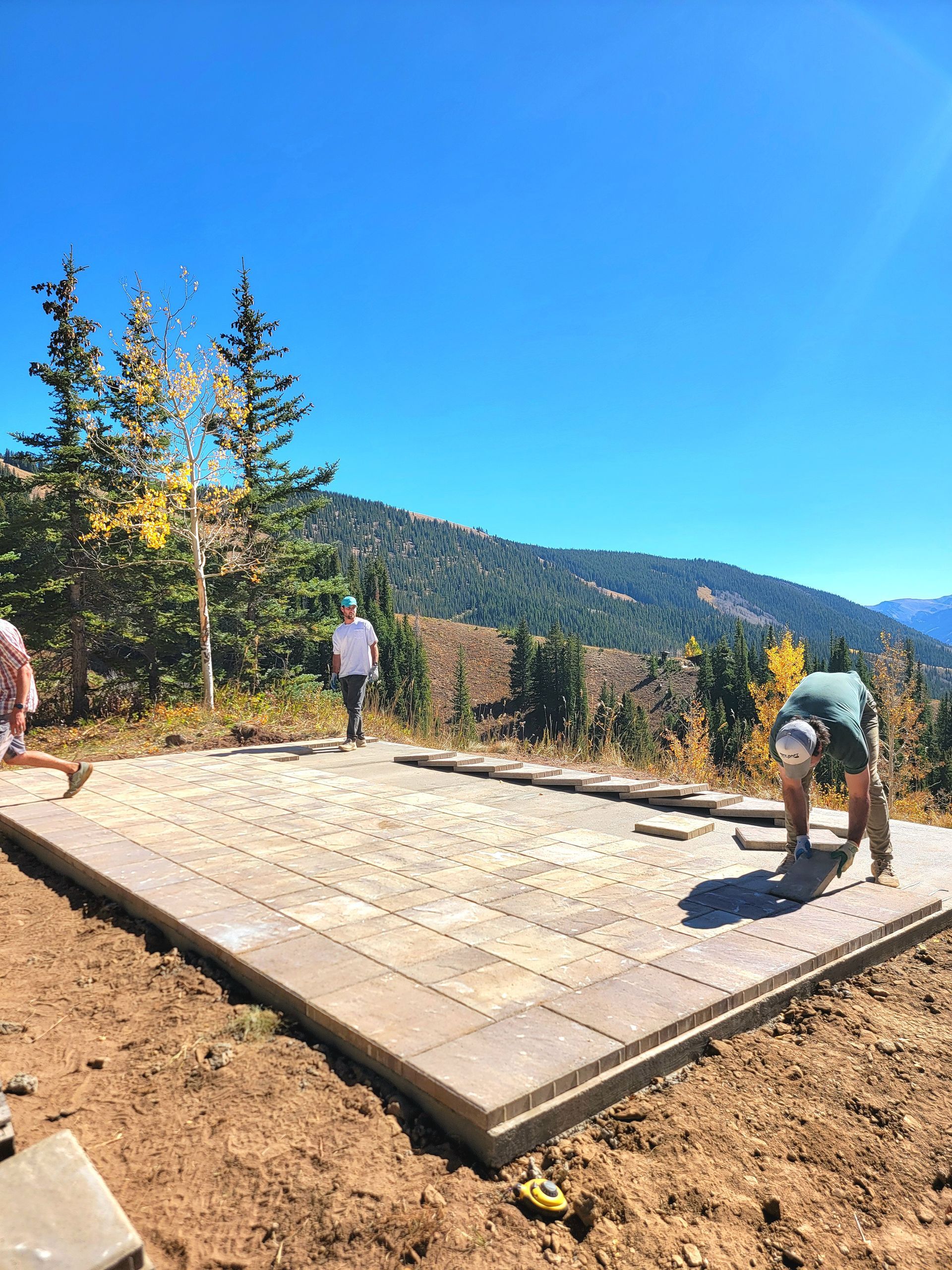 Workers laying stone pavers on a sunny outdoor patio with mountain views