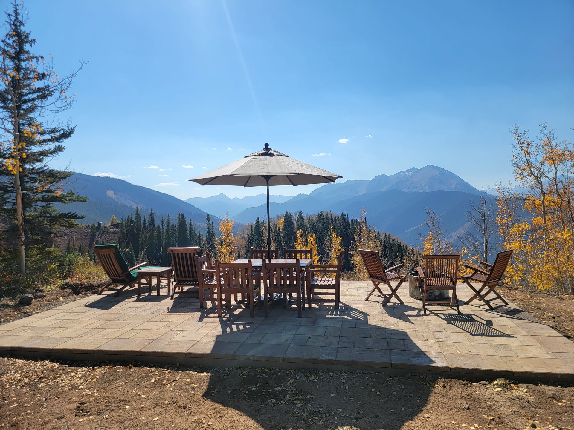Mountain patio with wooden tables and chairs under an umbrella, overlooking blue ridges and autumn trees.