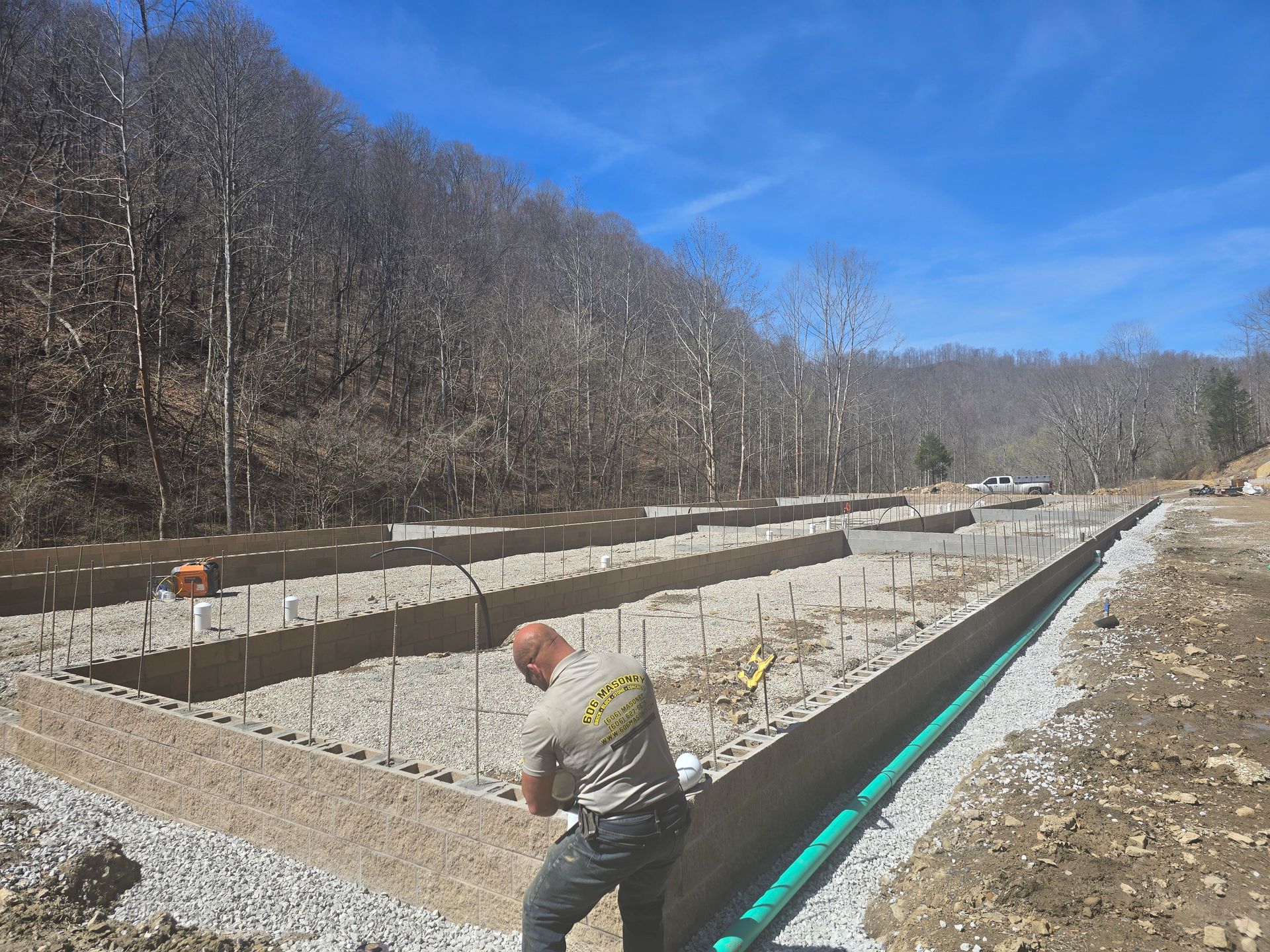 Worker installing drainage in a gravel-lined trench at a construction site beside a wooded hillside
