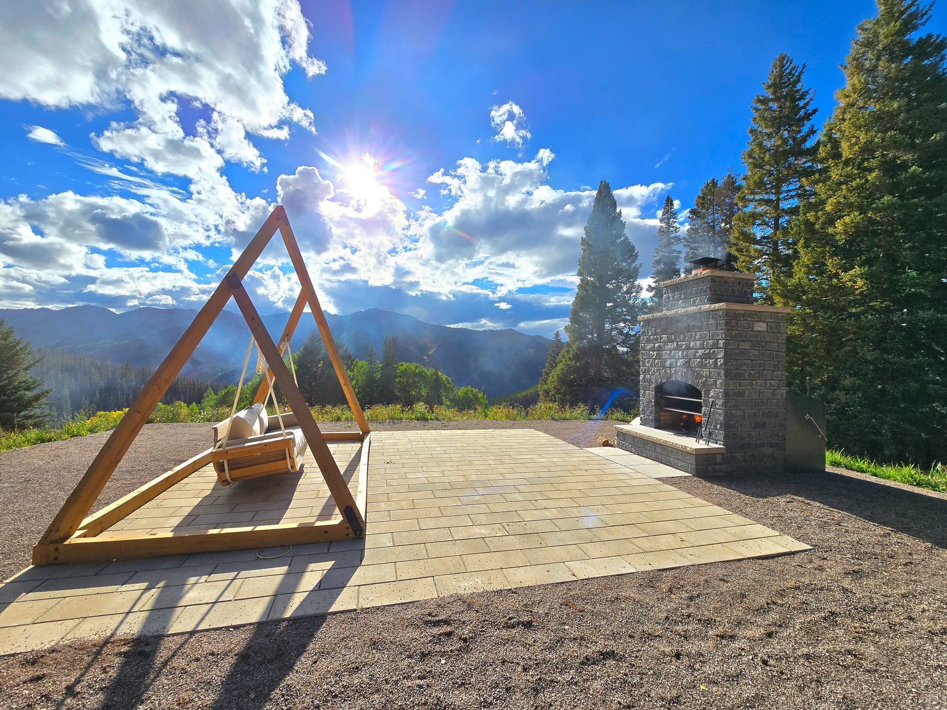 Sunlit mountain patio with wooden A-frame swing, fire pit, and forest view