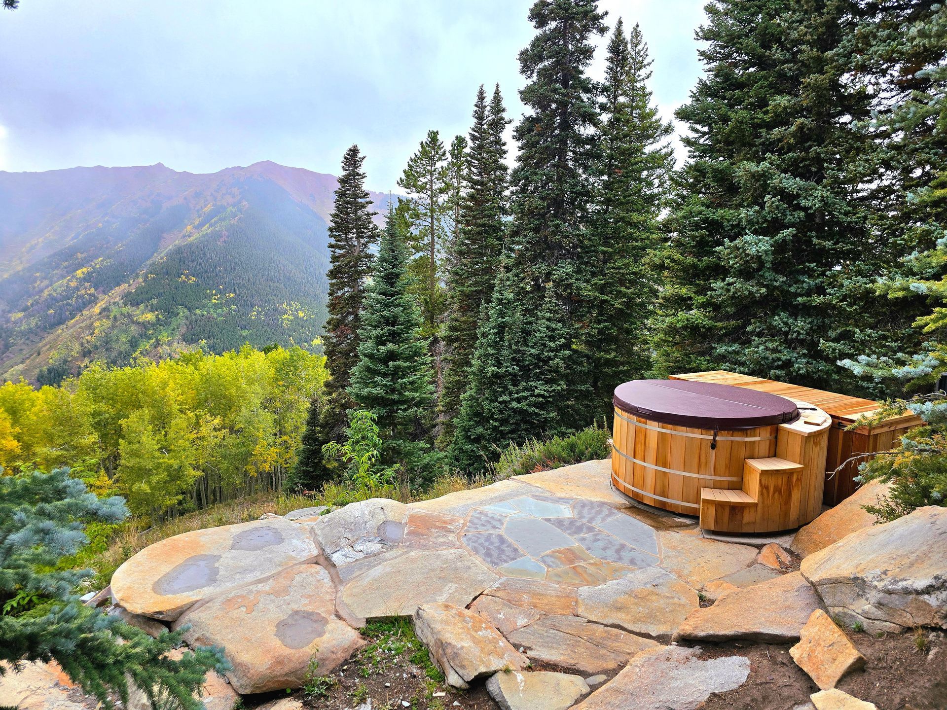 Mountain deck with a wooden hot tub overlooking pine trees and a valley at sunset