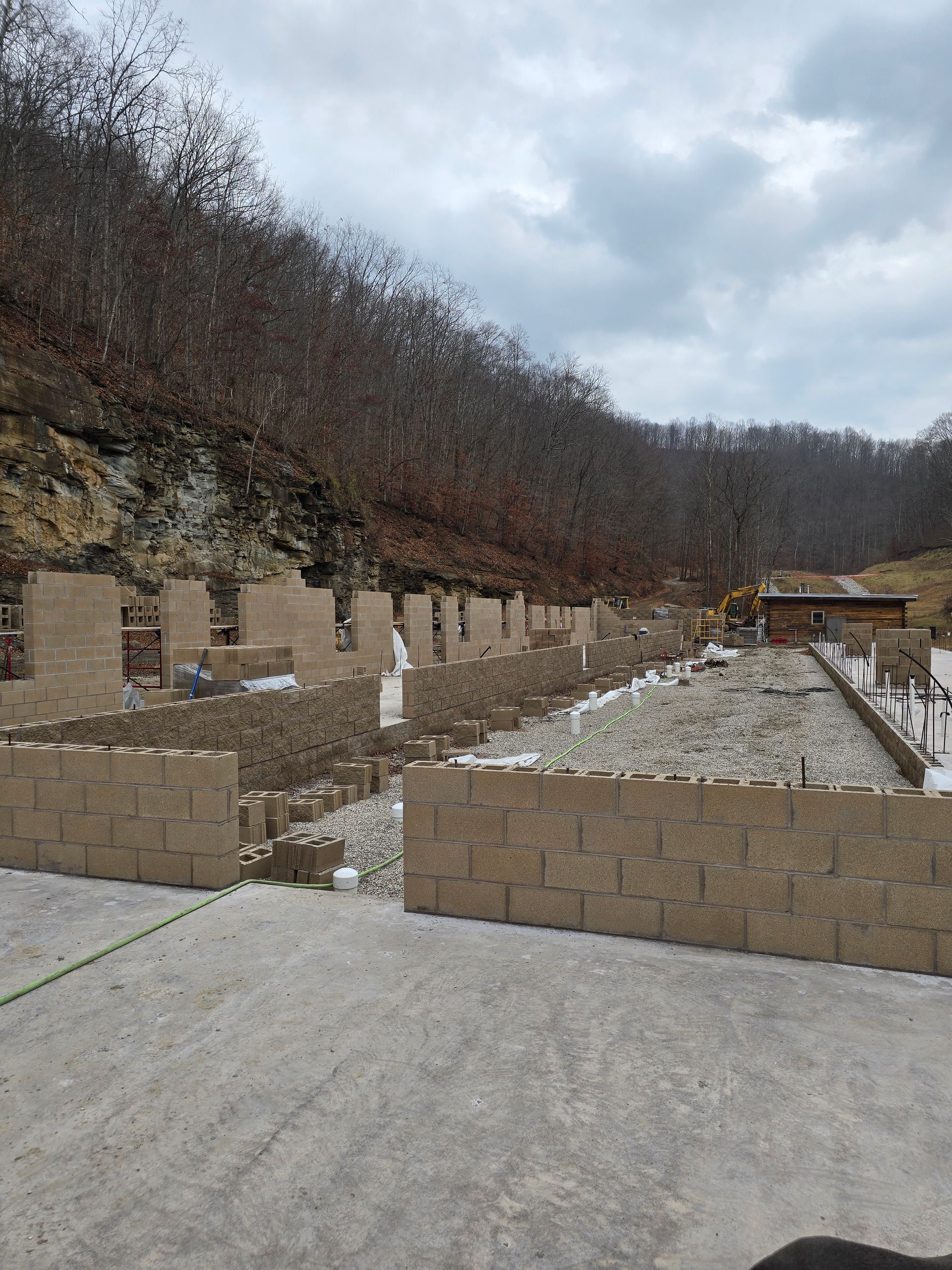 Construction site with concrete block walls under construction beside a wooded hillside and cloudy sky