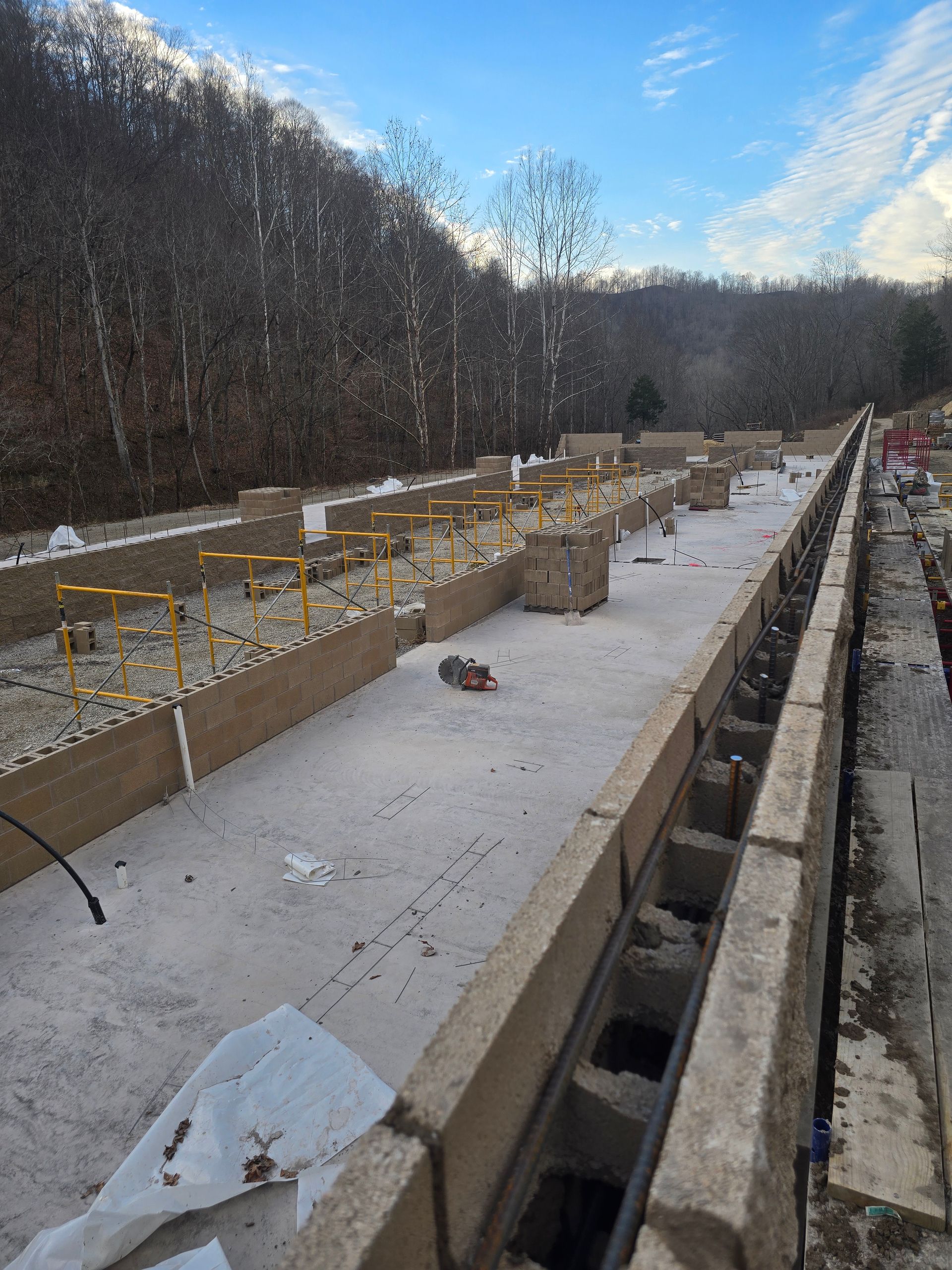 Snowy construction site with concrete foundation forms and yellow rebar beside a wooded hillside
