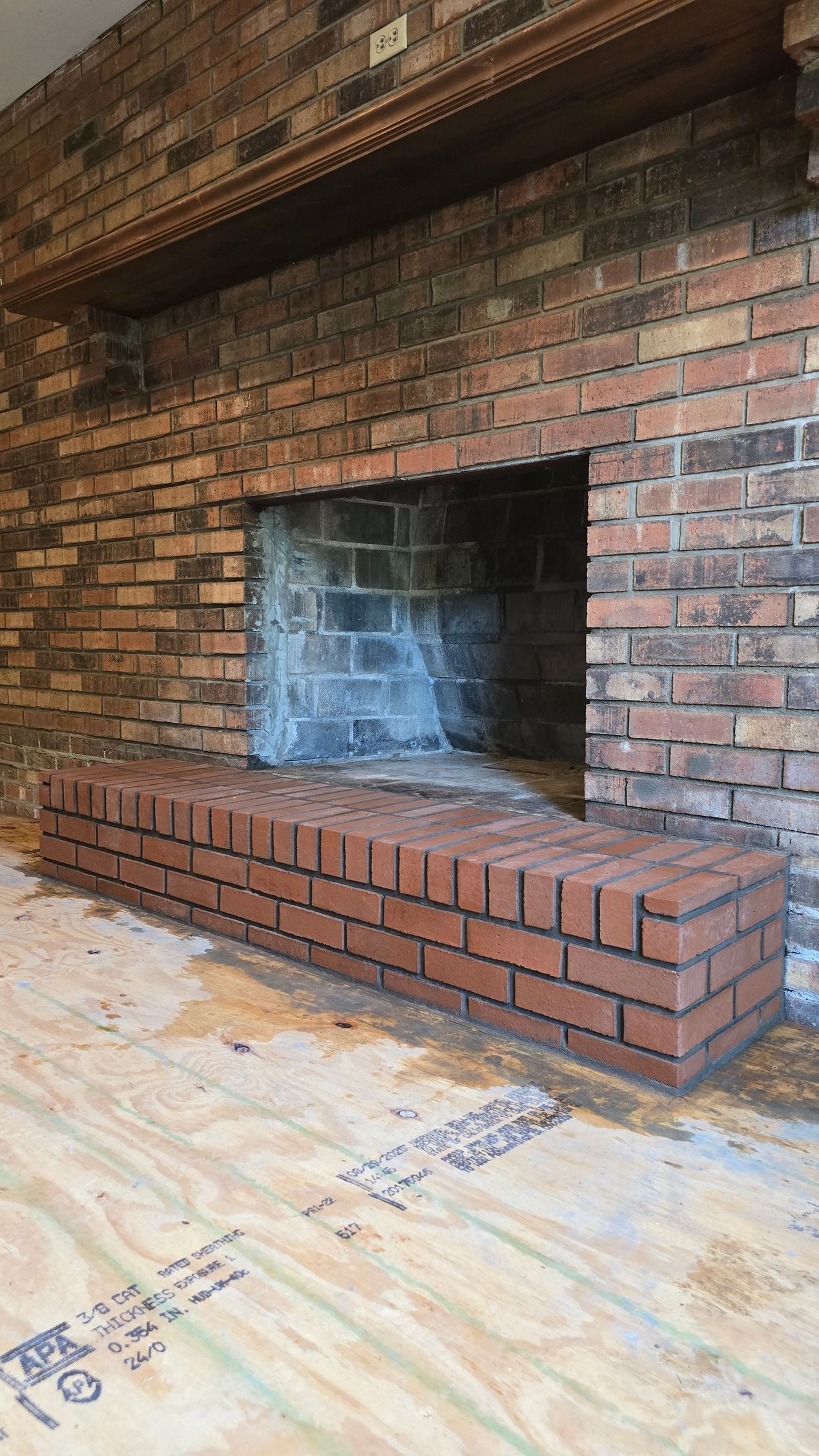 Brick fireplace with a raised hearth in an unfinished room with plywood flooring.