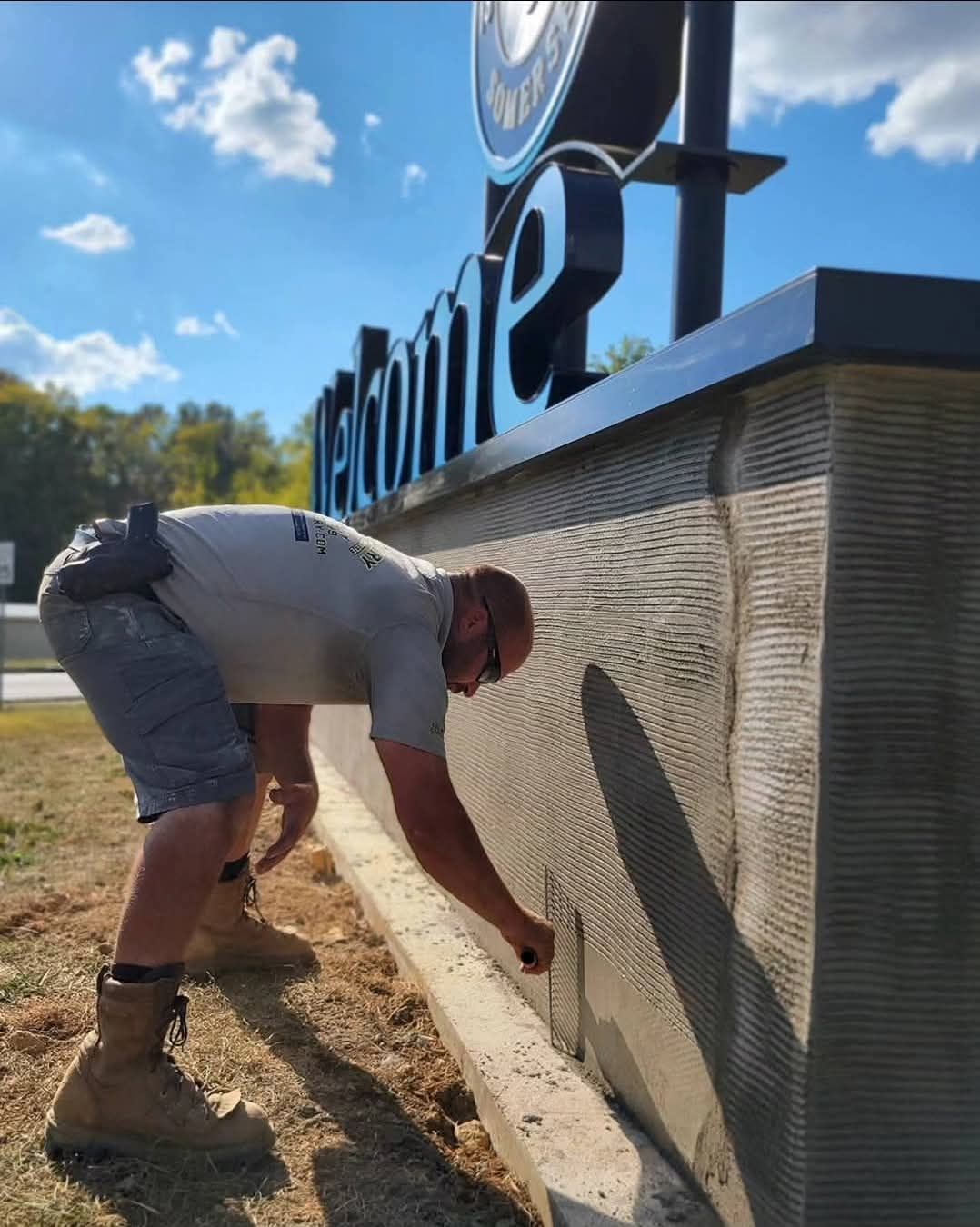 Worker bending over to spray paint a wall beside a large outdoor sign under a blue sky.