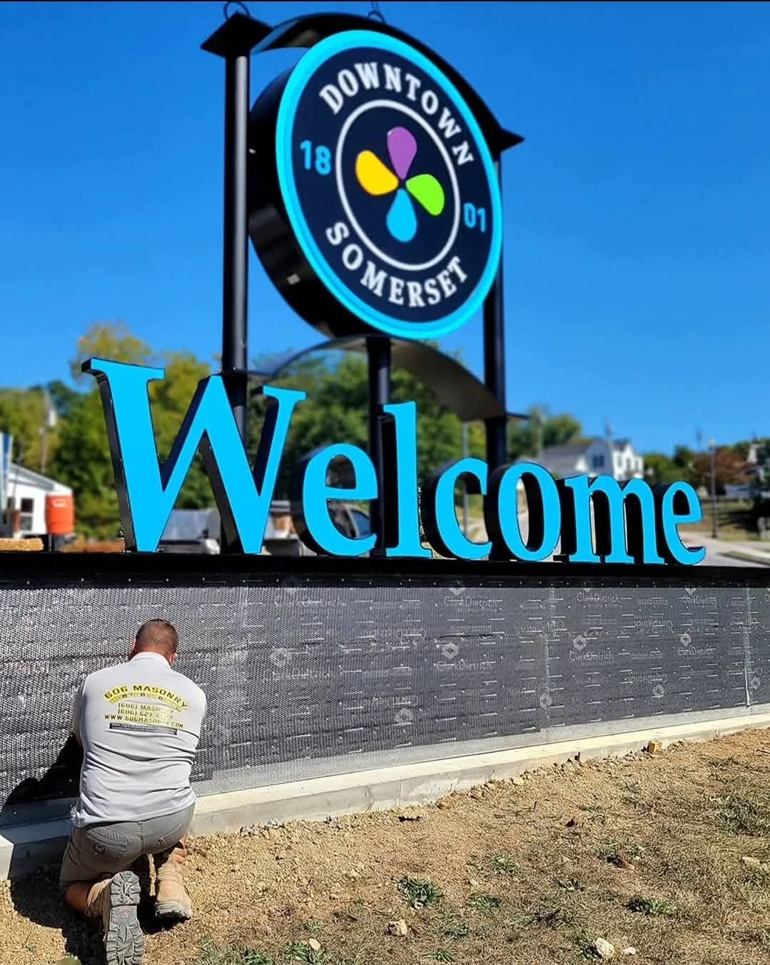 Downtown Somerset welcome sign with blue lettering and a person crouching at its base
