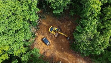 Aerial view of a yellow excavator and a car on a dirt clearing surrounded by dense green forest.