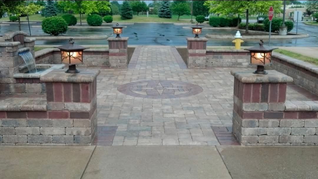 Stone walkway with lit pillars leading to a calm lake and green trees in the distance