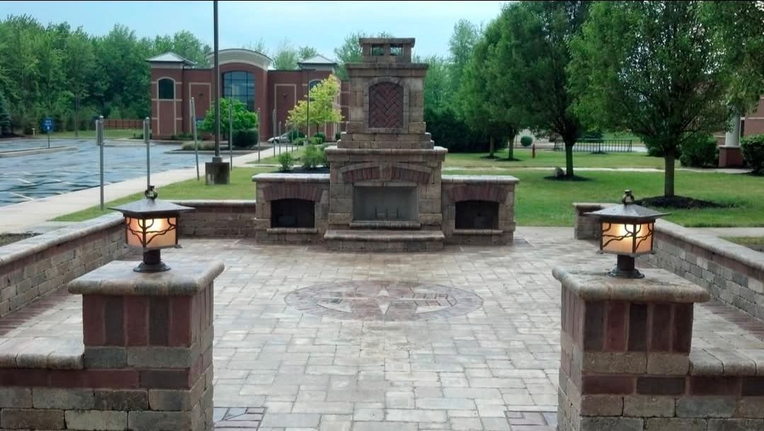 Brick memorial monument in a paved park, flanked by two lit lanterns and surrounded by trees