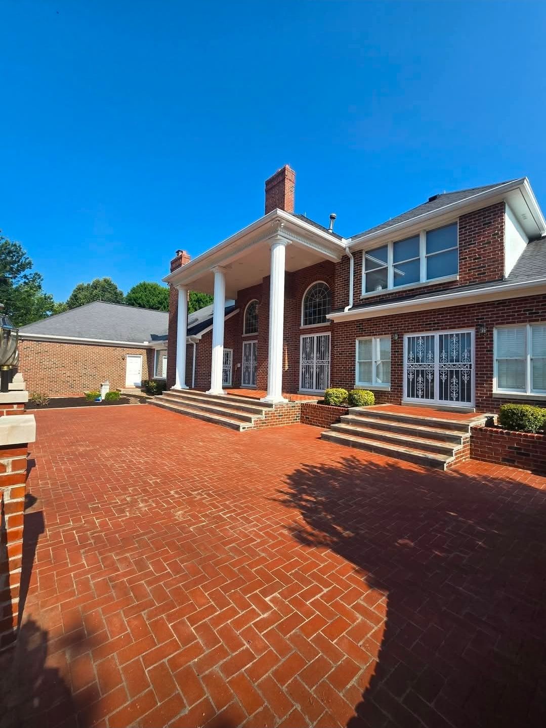 Brick driveway leading to a brick house with white columns and a covered front porch under a clear blue sky