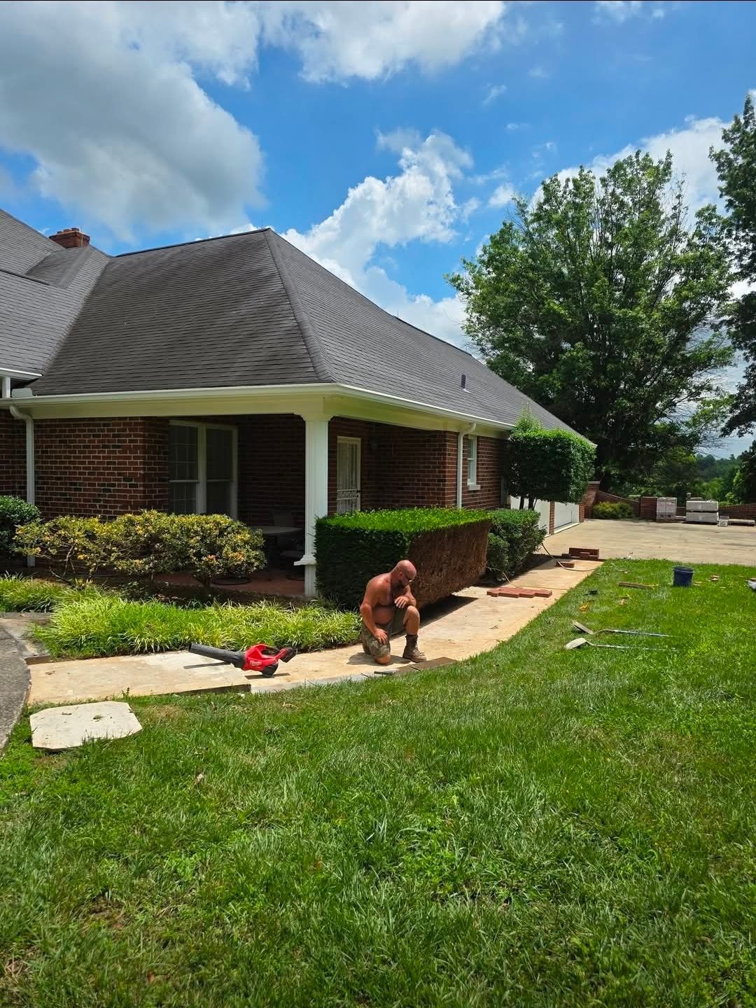 House with gray roof and brick porch in a green yard, with a person kneeling near the walkway under a blue sky