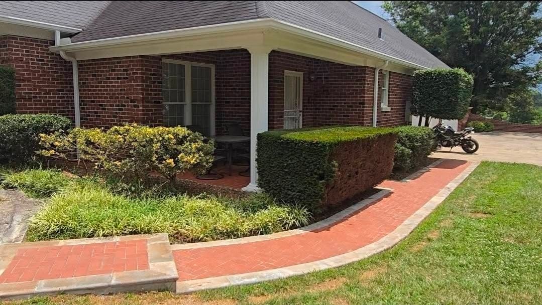 Brick house corner with white columns, trimmed hedges, and a red walkway beside a green lawn