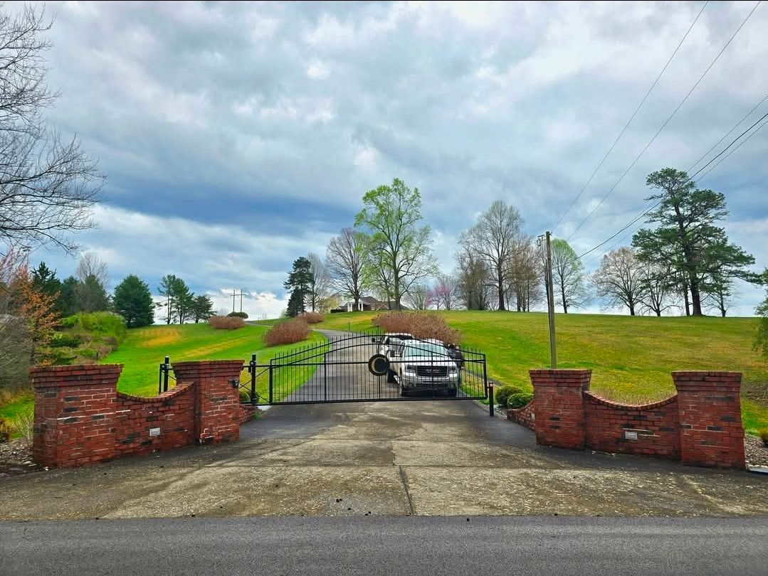 Driveway entrance with open gate, brick pillars, and a car parked on a grassy hill under cloudy skies