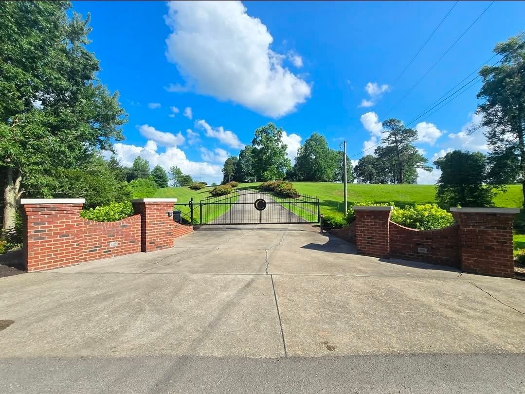 Closed iron gate at the end of a concrete driveway, with brick pillars and green hills under a blue sky