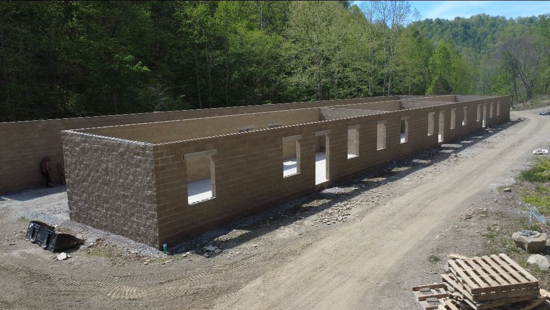 Long brick building under construction along a dirt road in a wooded area
