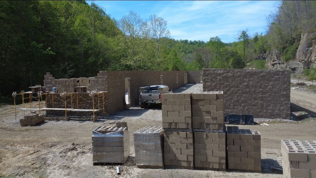 Stacked concrete blocks at a construction site beside stone walls and trees under a blue sky