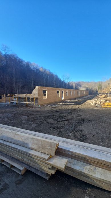 Partially built wooden house on a dirt lot with lumber stacks in the foreground and wooded hills behind