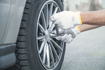 Gloved mechanic uses a lug wrench on a shiny alloy car wheel and tire, conveying careful, hands-on vehicle maintenance.