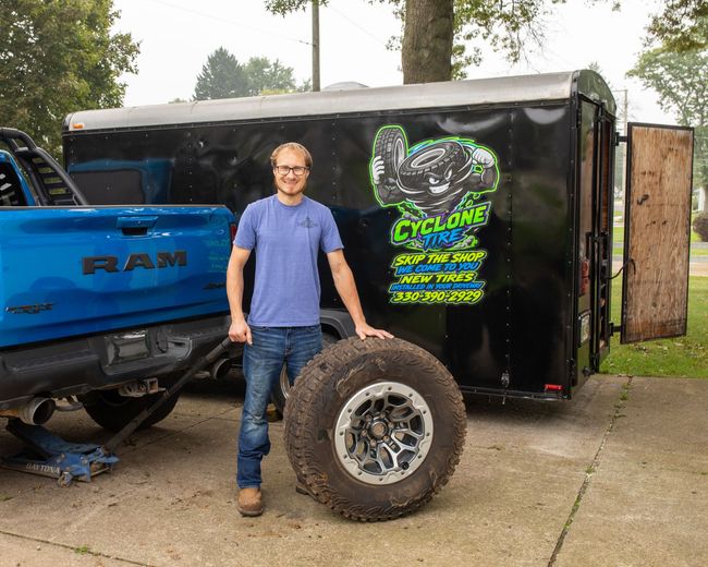 Black trailer featuring a colorful Cyclone Tire cartoon mascot holding tires, with bold promotional text and phone number, parked outdoors.