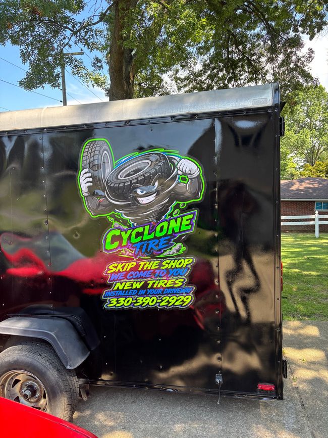 Black trailer featuring a colorful Cyclone Tire cartoon mascot holding tires, with bold promotional text and phone number, parked outdoors.