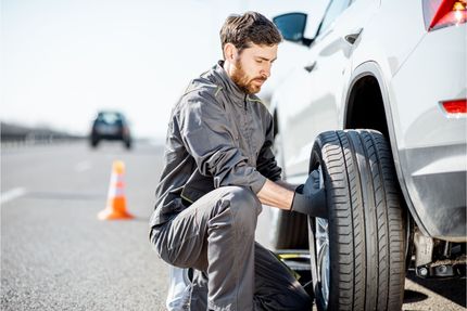 Mechanic in gray coveralls kneels beside a white SUV, inspecting a removed tire on a road shoulder near an orange safety cone.