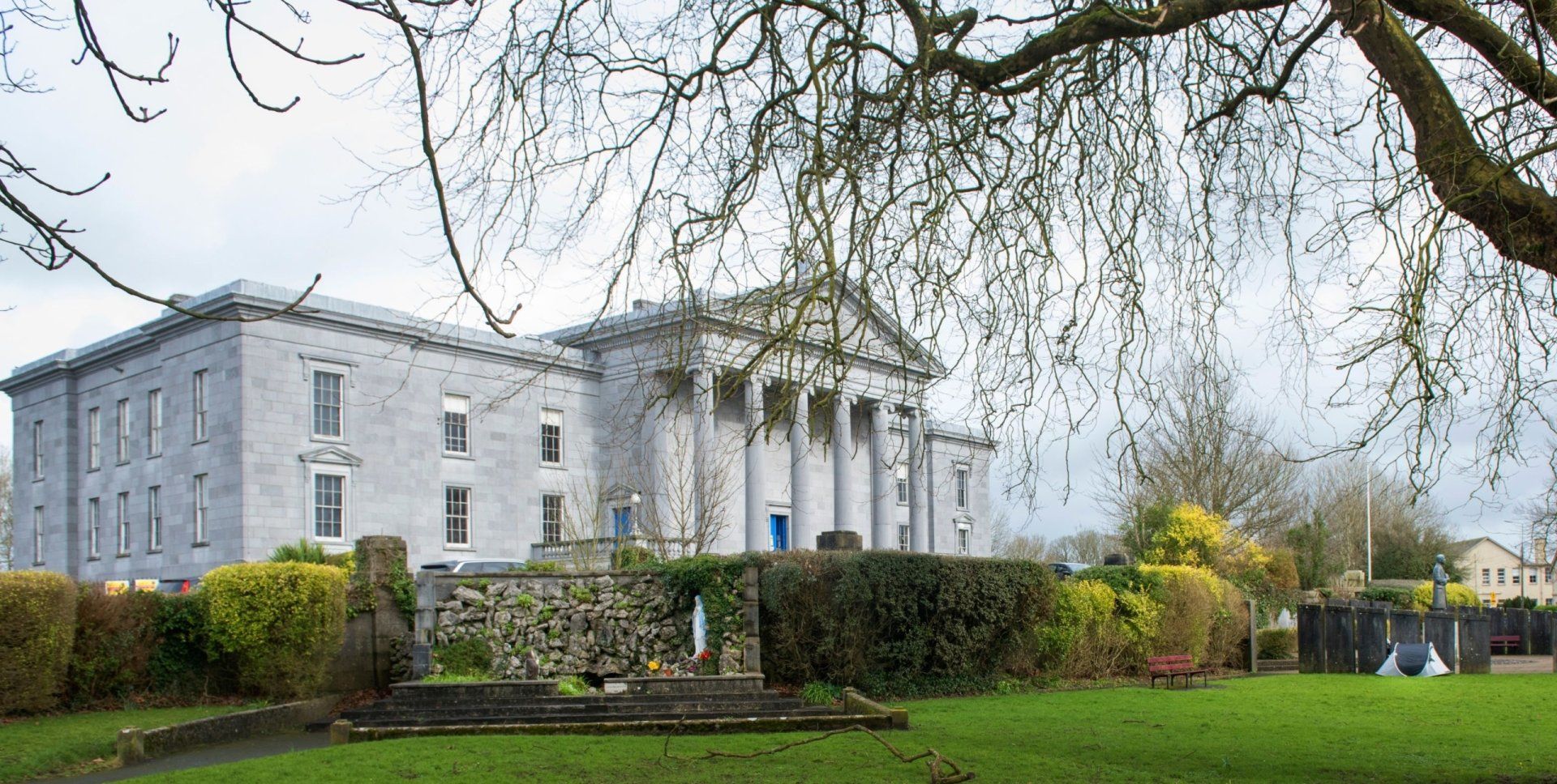 Large white mansion with columns beside a manicured green lawn and hedges under bare tree branches