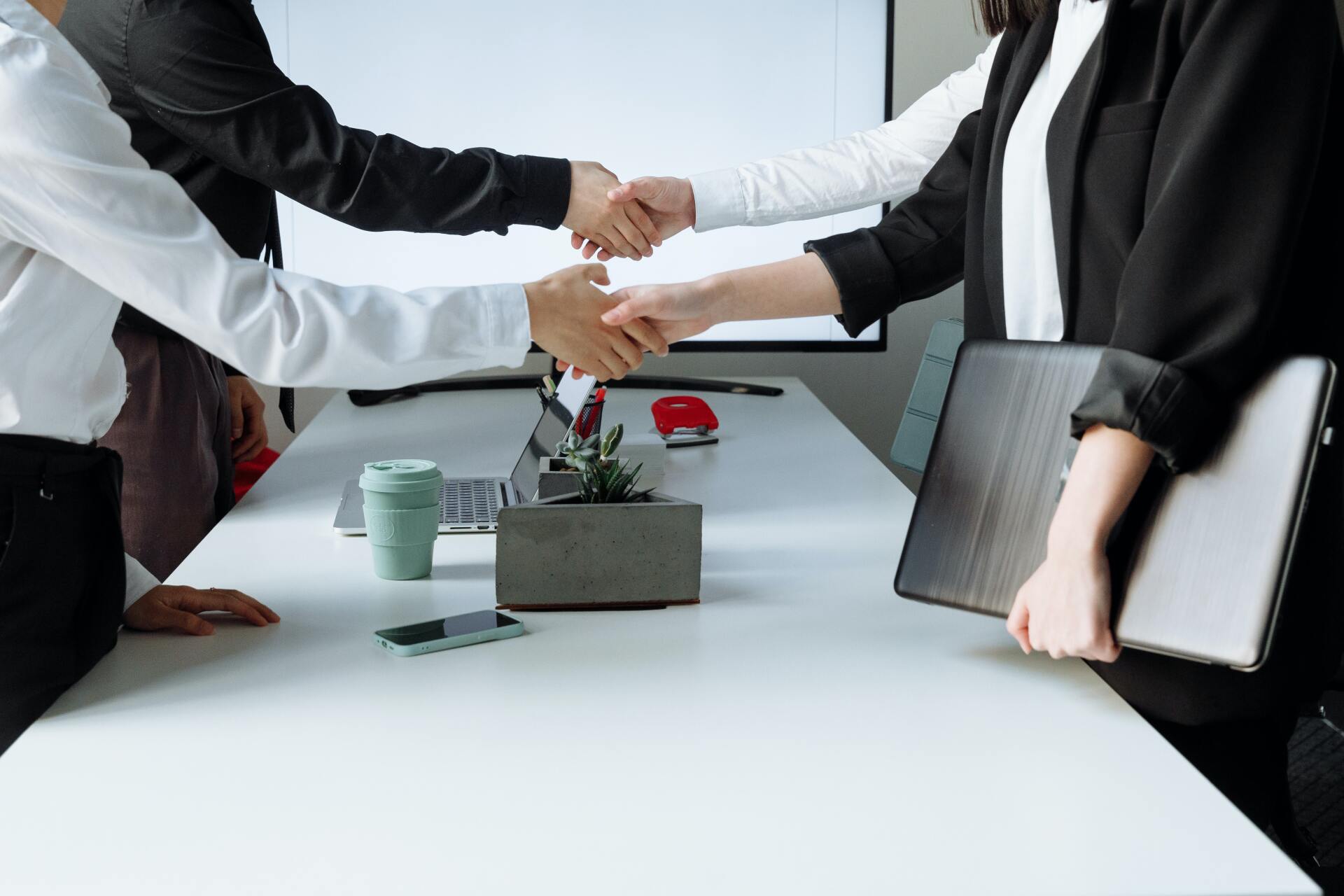 Business people shaking hands across a conference table with laptop, coffee cups, and flowers