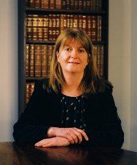 Woman seated at a table in front of bookshelves, wearing a black jacket and slight smile.