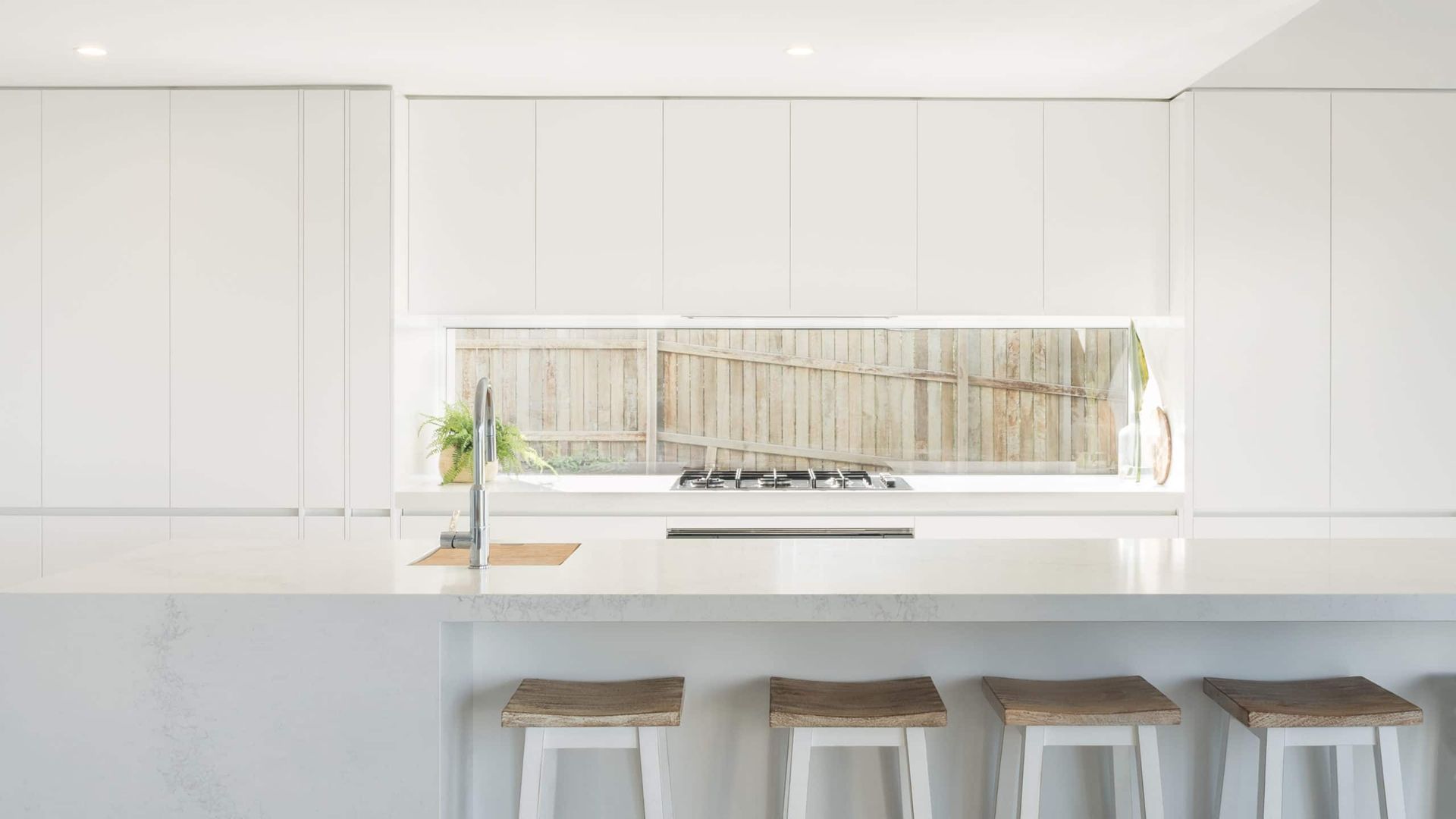 Modern White Kitchen With a Window Behind the Cooktop and Wooden Stools at a Counter — Akyrah Kitchens in Gympie, QLD