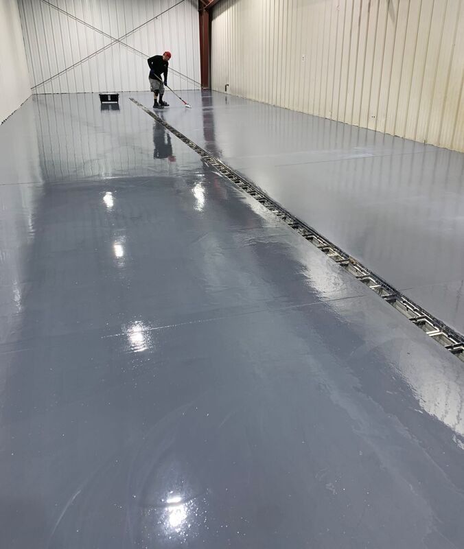 Person using a floor machine on a glossy gray industrial floor inside a warehouse-like room