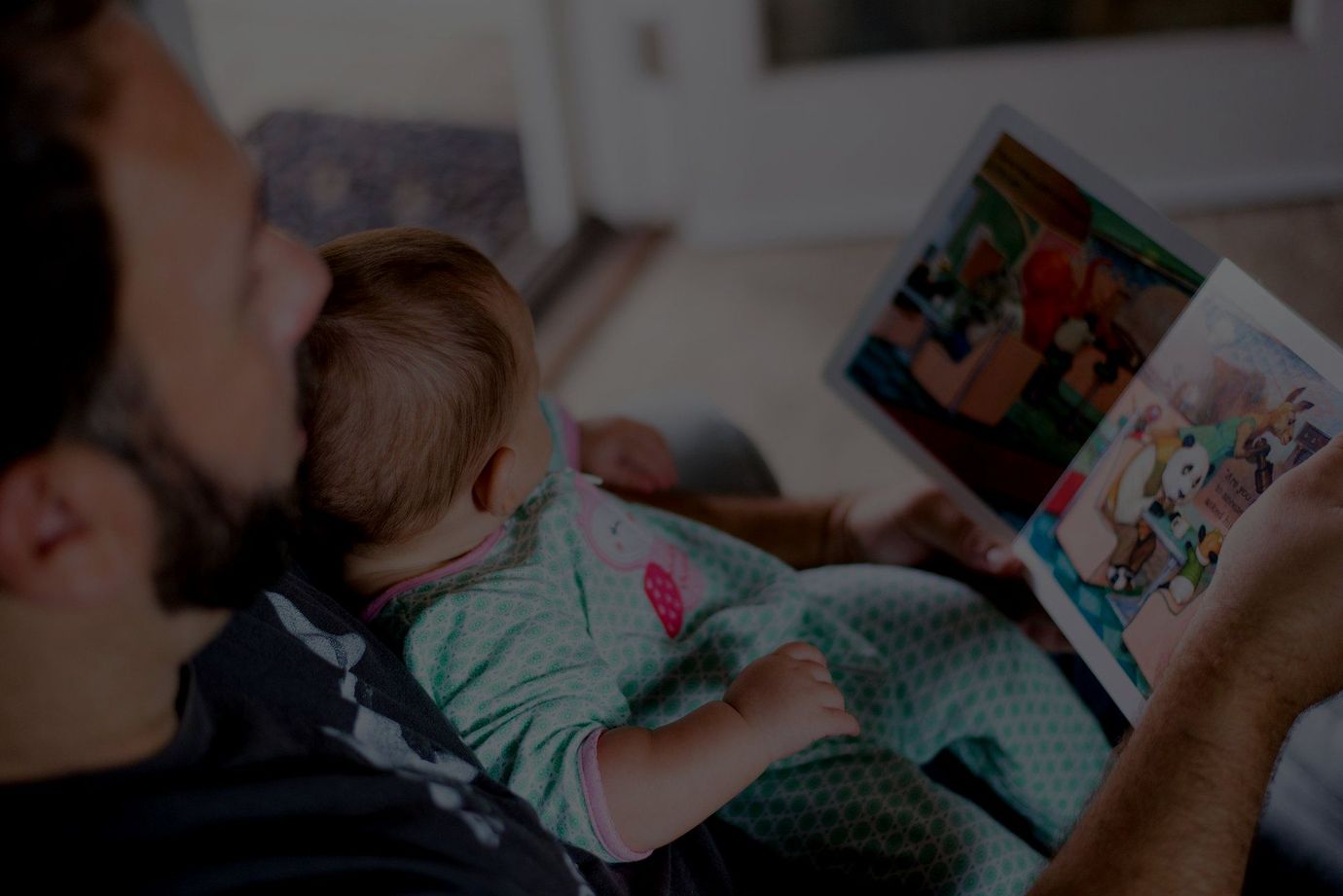 A father reading a picture book to his infant in a chair.