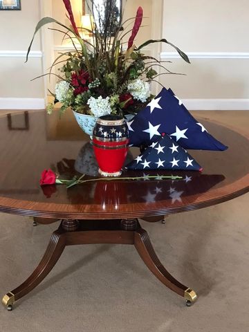 Floral arrangement and folded American flag displayed on a polished wooden table with a red rose in front.