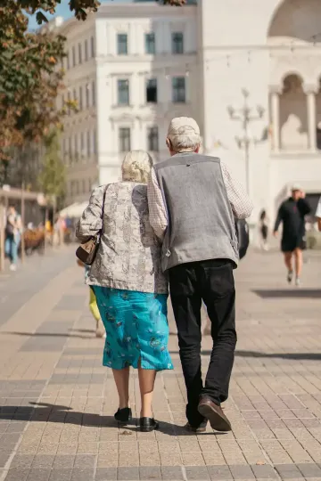 Two elderly people walking arm in arm on a city sidewalk, seen from behind