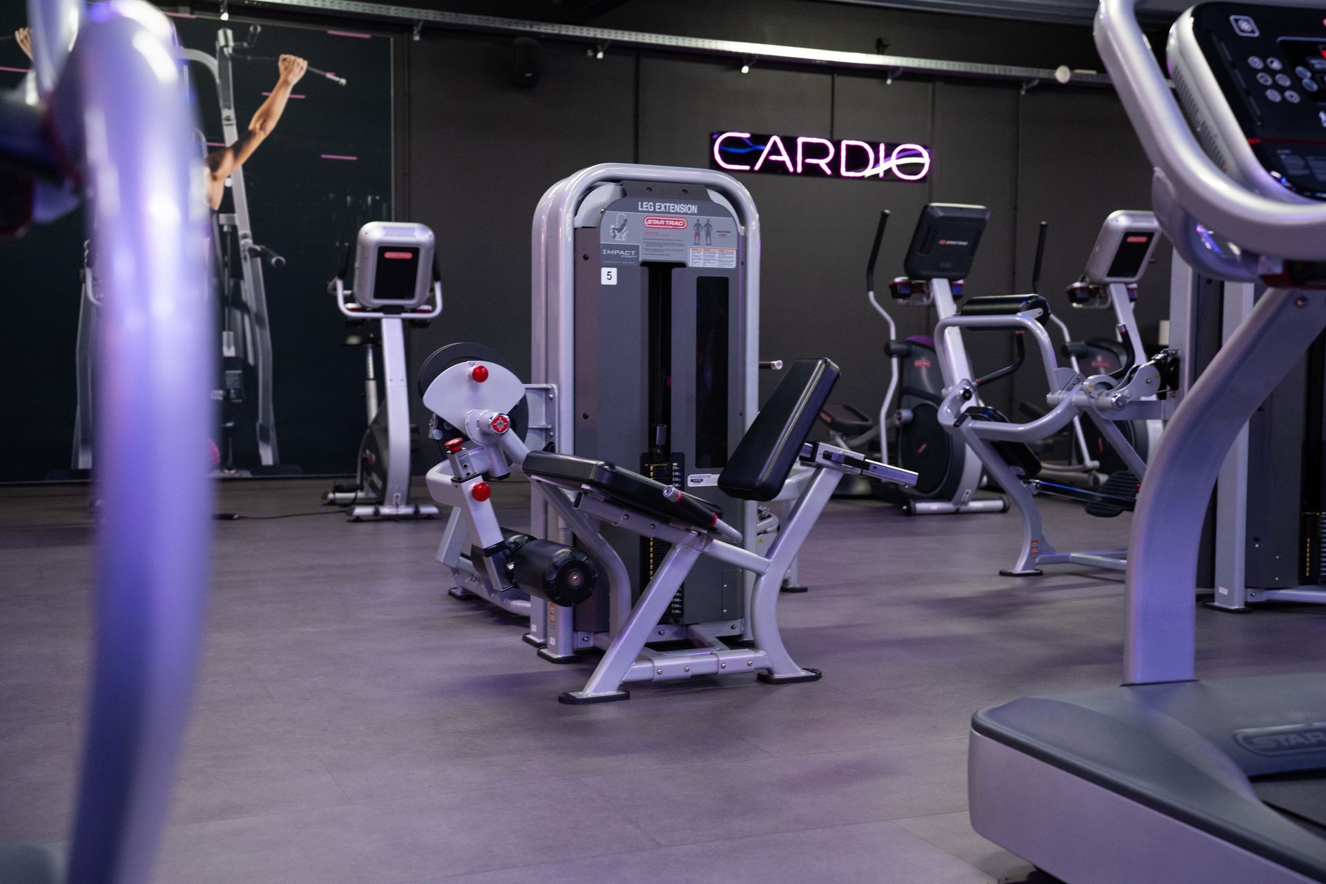 Lege sportschool met hometrainers, loopbanden en krachtapparaten onder paarse verlichting.