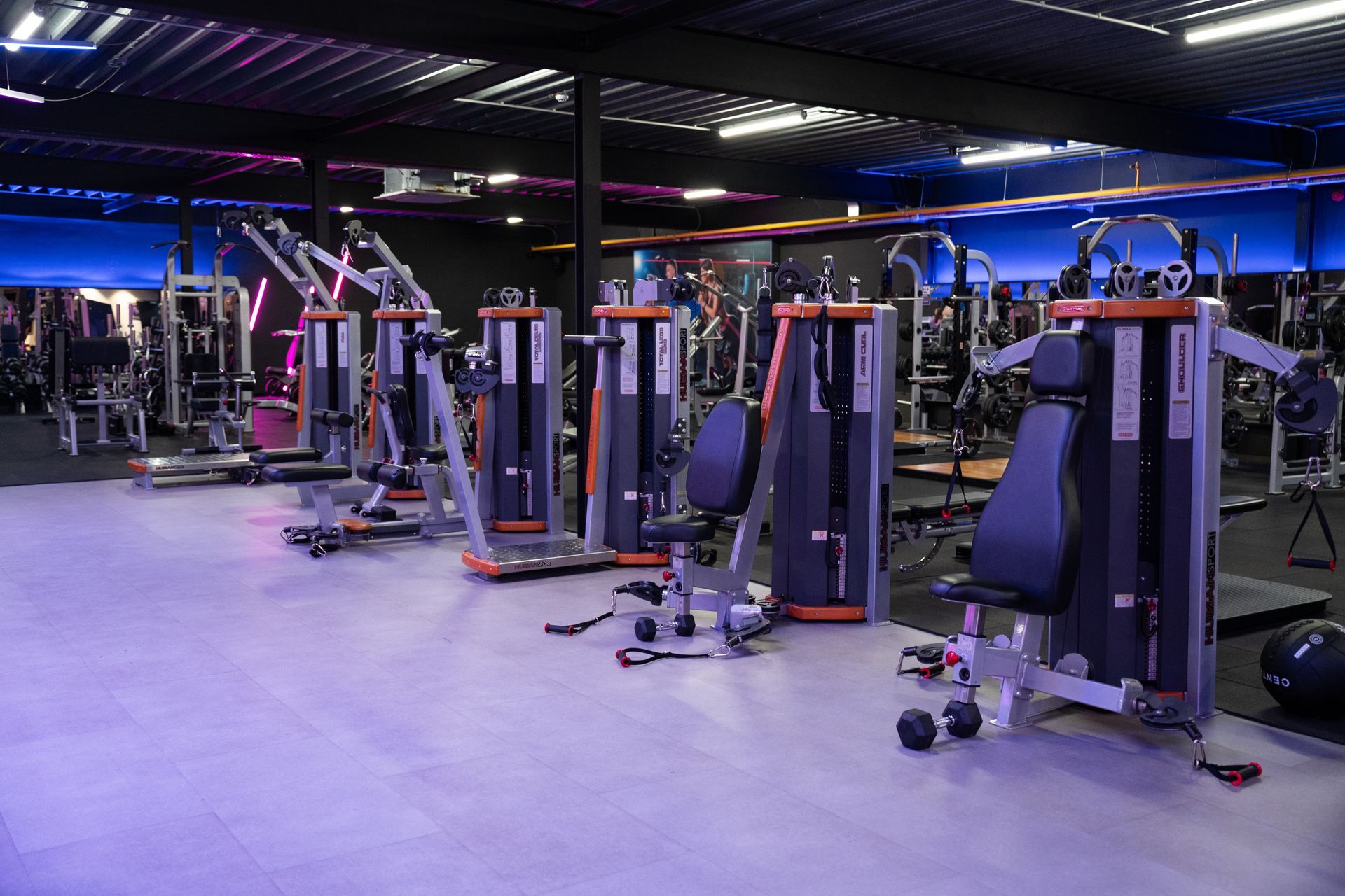 Modern fitnesscentrum met rijen krachtapparaten en paarse verlichting.