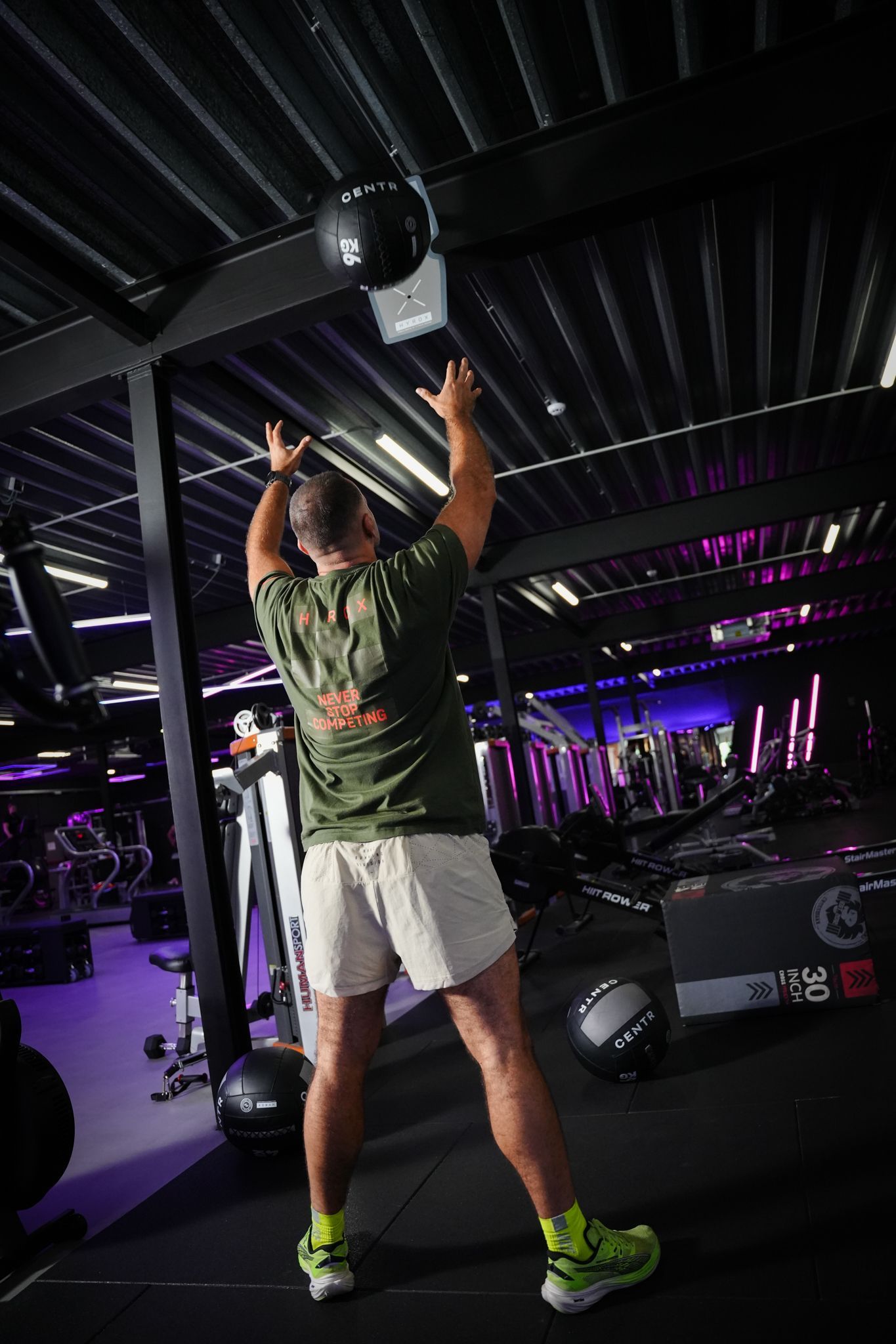 Een persoon tilt een kettlebell boven zijn hoofd in een sportschool met paarse verlichting en fitnessapparatuur.