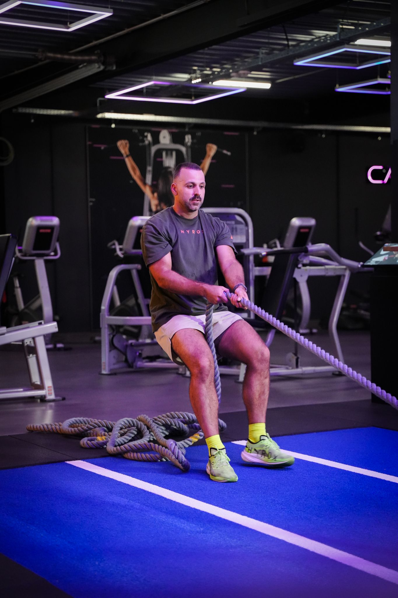 Een man traint met battle ropes in een sportschool, staand op een blauwe vloer vlakbij loopbanden.