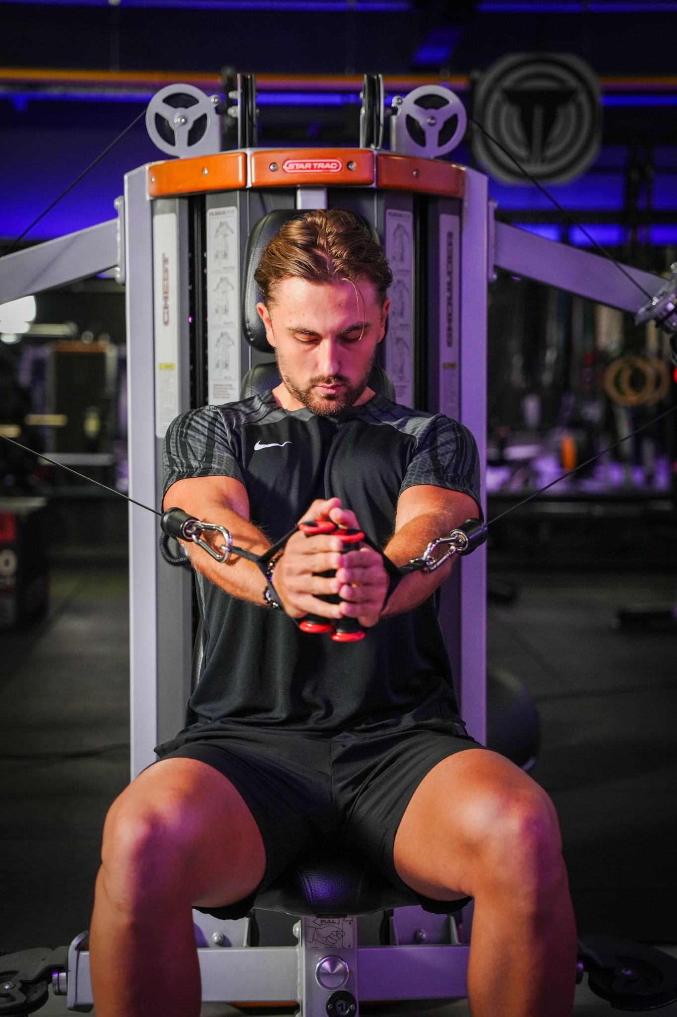 Persoon die een chest fly-machine in een sportschool gebruikt, met de blik naar beneden gericht, verlicht door paars licht.