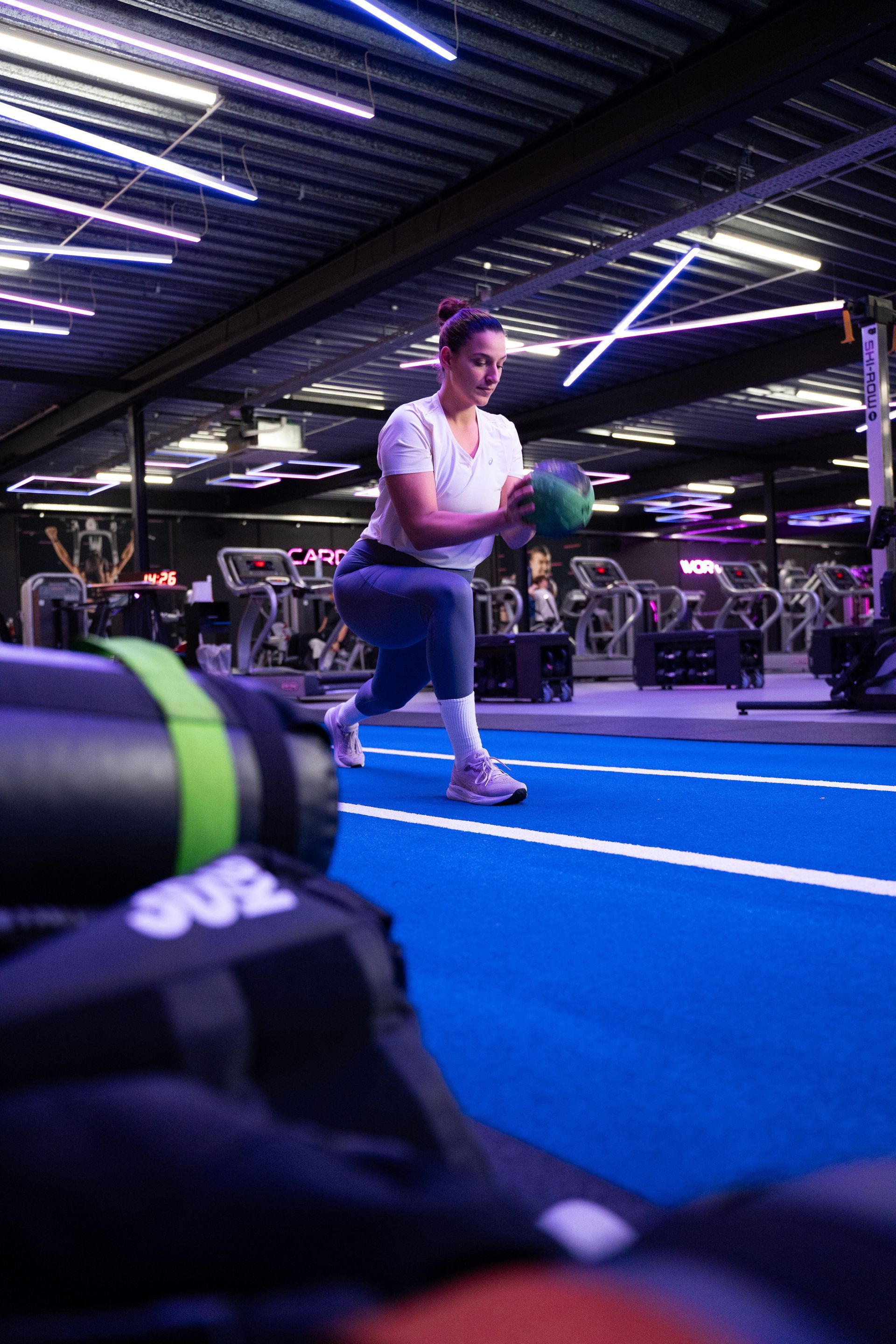 Persoon stretcht op blauwe atletiekbaan in sportschool met fitnessapparatuur en neonverlichting.