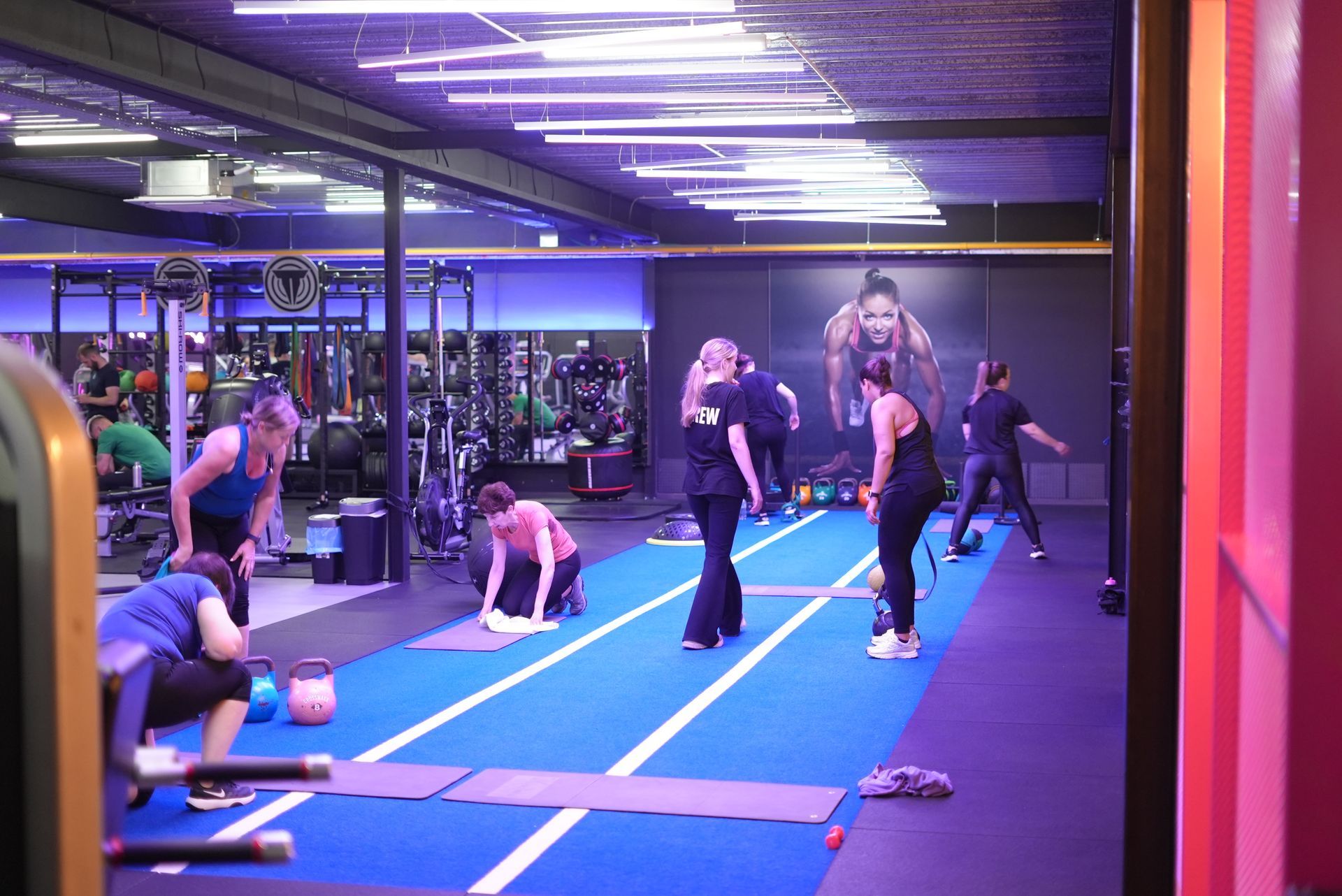 Mensen sporten op blauwe matten in een sportschool met spiegels en fel verlichte plafondlampen.