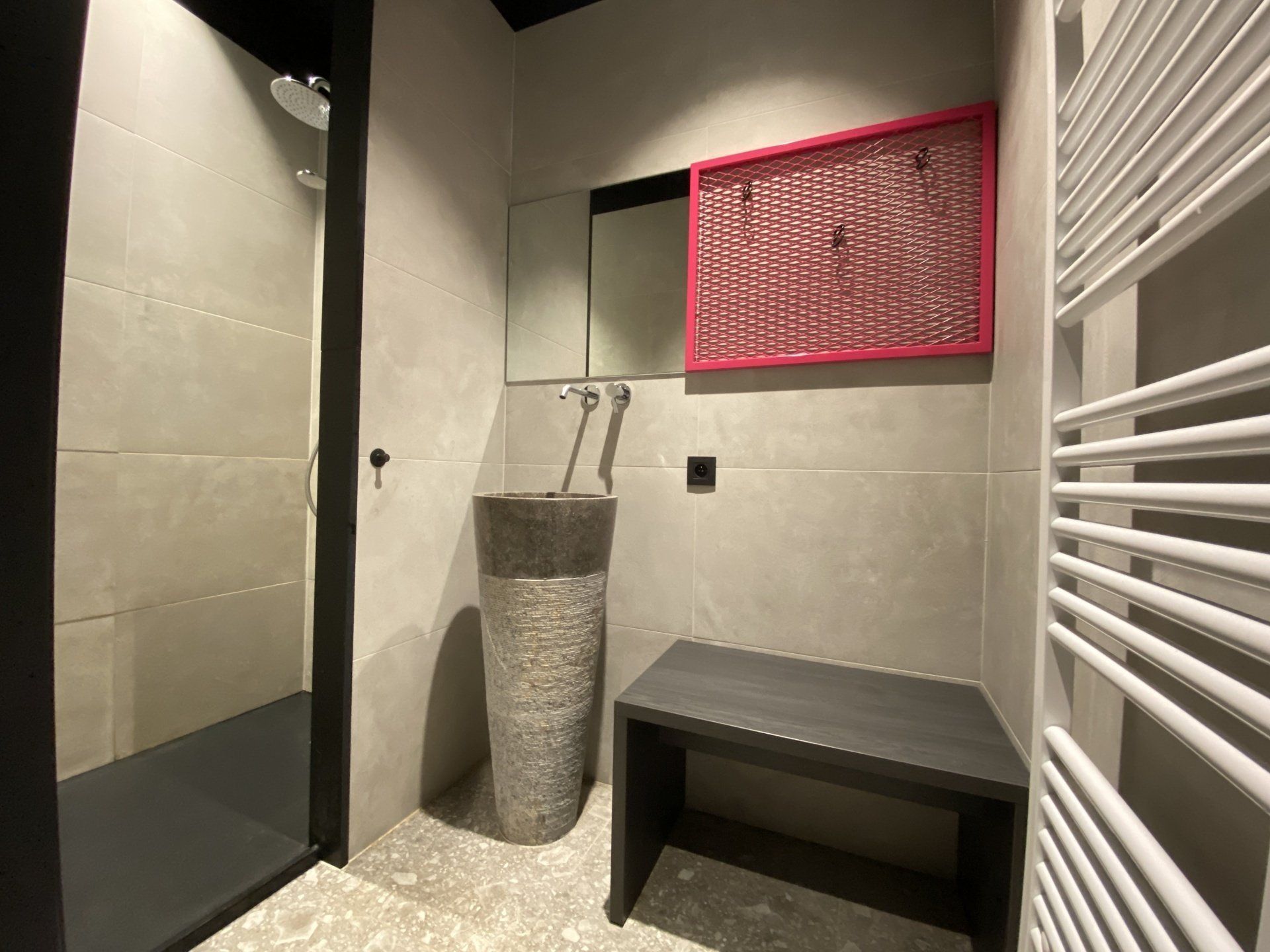 Moderne grijze badkamer met een stenen wastafel, zitbank, zwarte douche en een spiegel met rode lijst op betegelde muren.