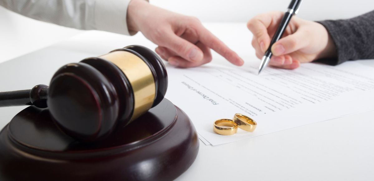 Judge’s gavel beside hands signing legal documents with wedding rings on a white desk