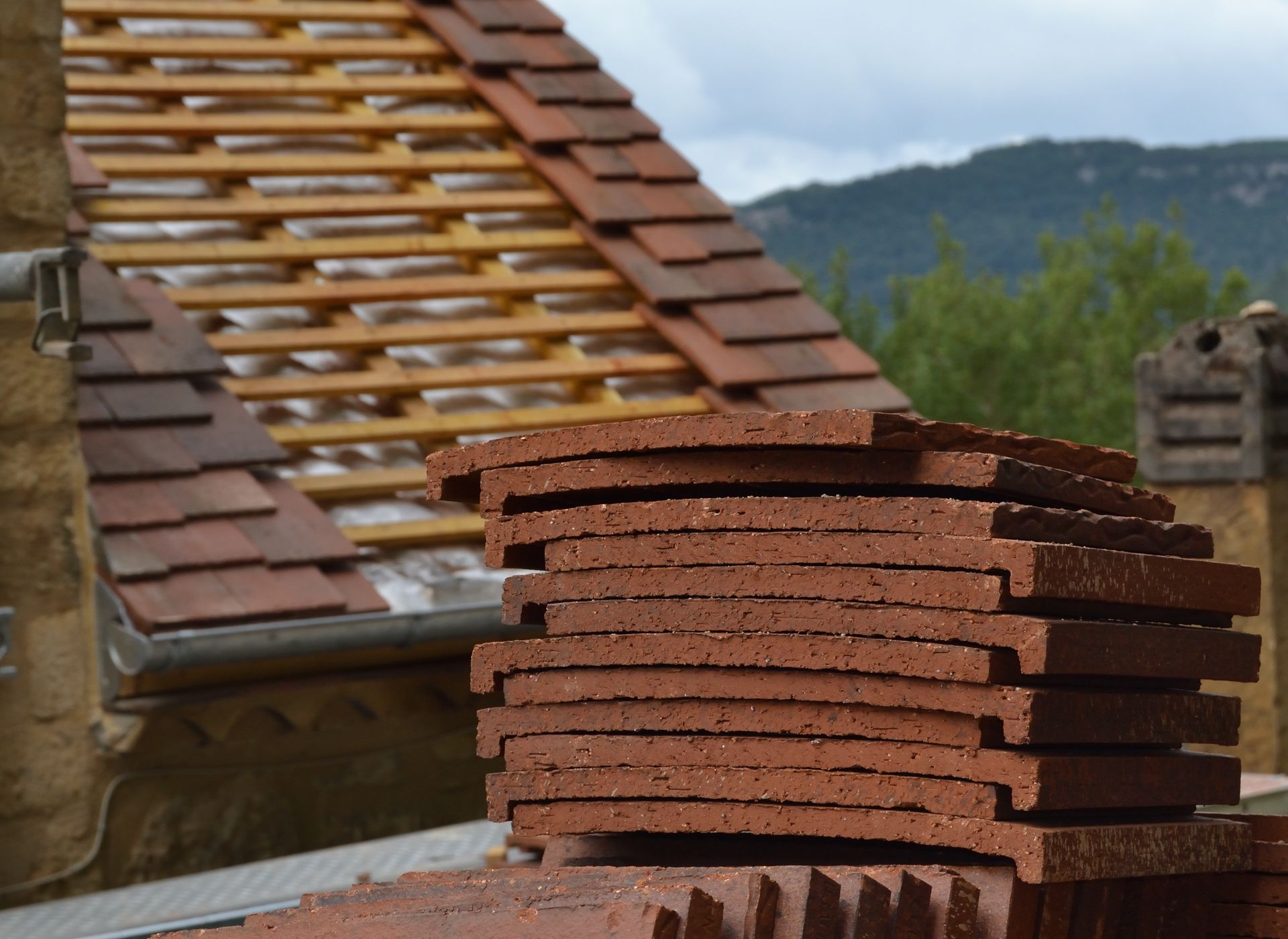 Stacked red roof tiles in the foreground with a tiled rooftop and cloudy mountains behind.