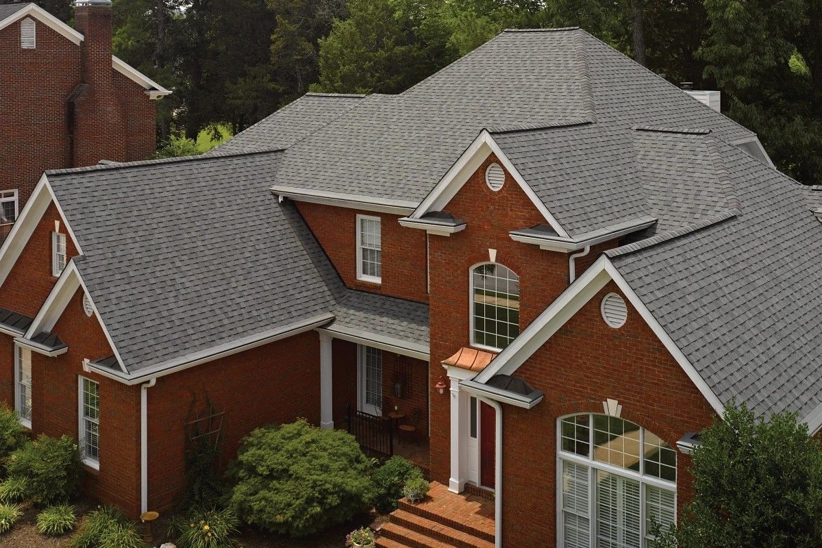 Aerial view of a large red brick house with gray shingle roof and white-trimmed windows surrounded by trees