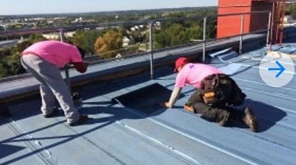 Two workers in pink shirts installing roofing on a flat rooftop with city view in the background