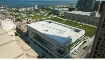 Aerial view of a modern white building near a waterfront, with green lawns and nearby high-rises.
