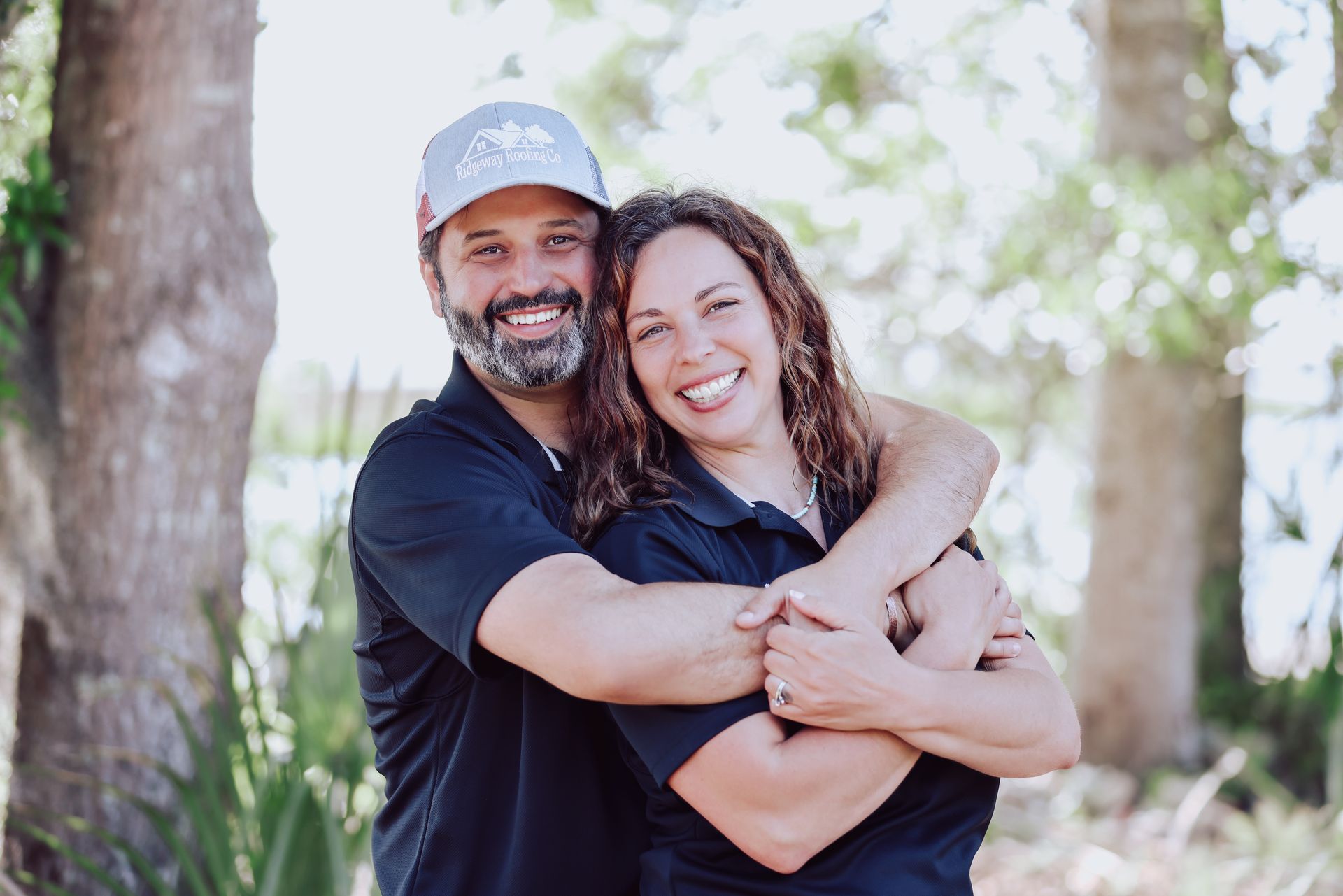 Couple embracing outdoors, smiling at the camera, trees in the background.