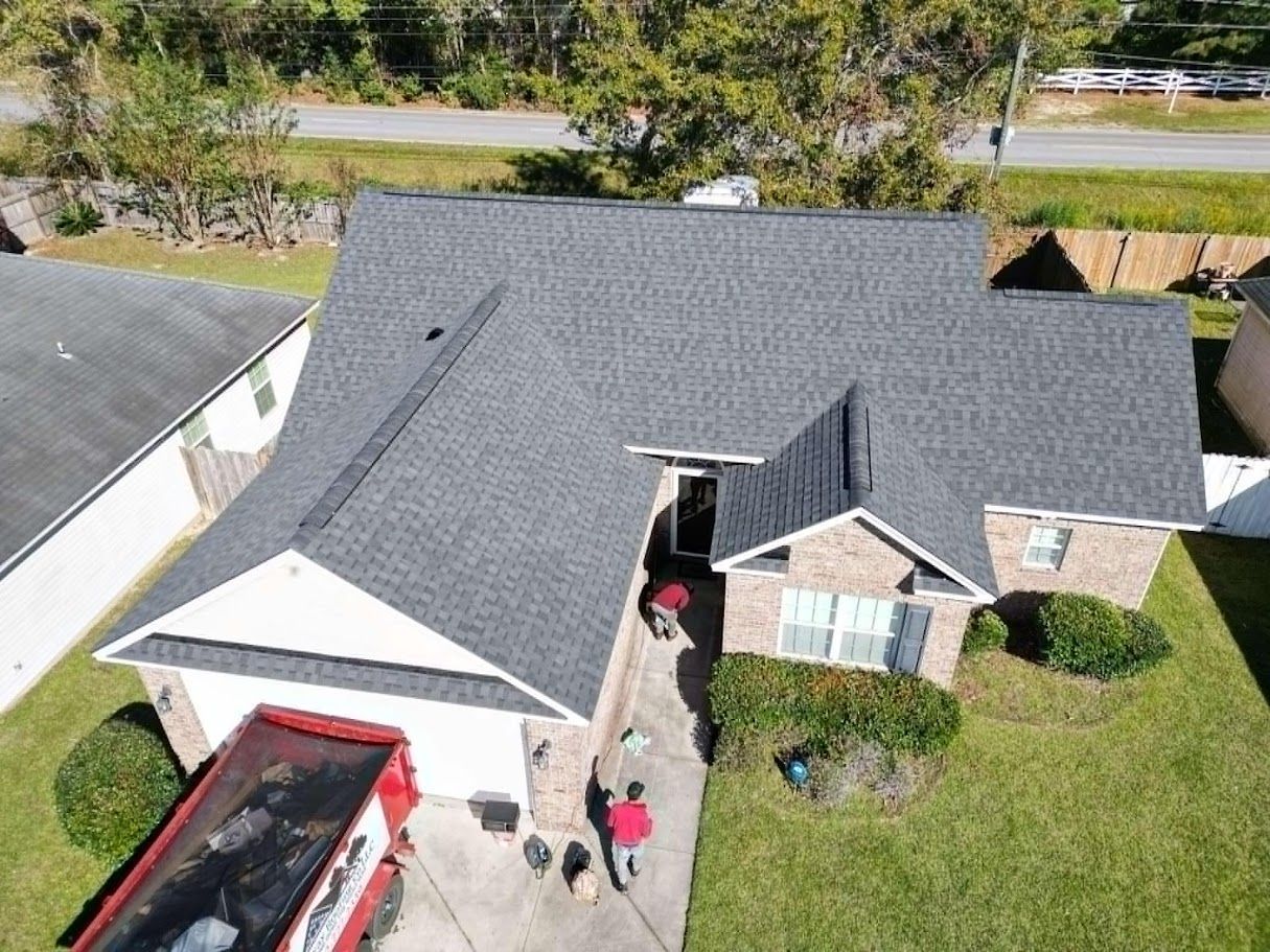 Aerial view of a house with a newly installed gray roof; workers are near a dumpster in the driveway.