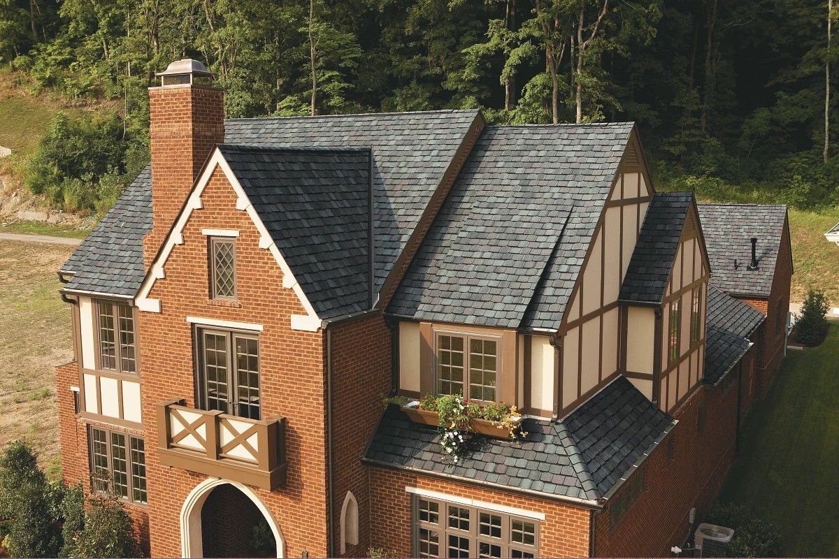 Brick-and-timber house with a steep gray roof and chimney, surrounded by trees and greenery