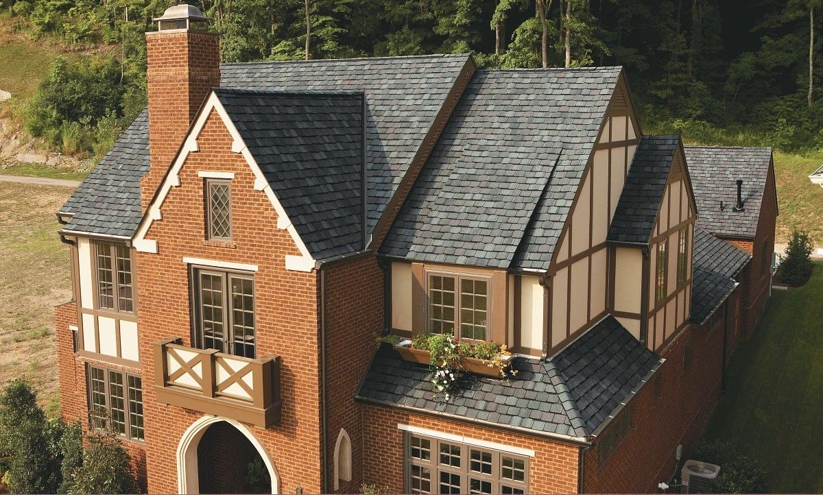 A red-brick Tudor-style house with a steep slate roof, chimney, and arched doorway amid greenery.